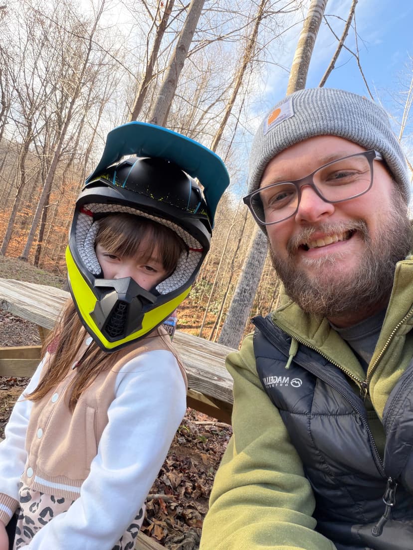 Child wearing a helmet with an adult, smiling outdoors in a forest setting.
